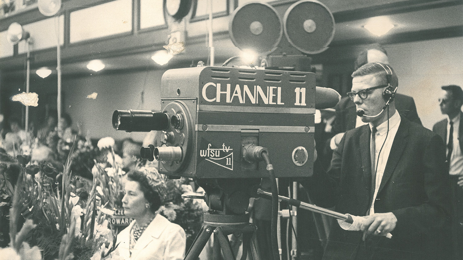 a historic shot of a young man on a WFSU-TV camera at the Florida Legislature. A woman in a hat sits nearby behind flowers.