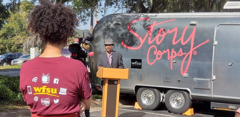 woman on camera recording man speaking at podium in front of story corps silver airstream