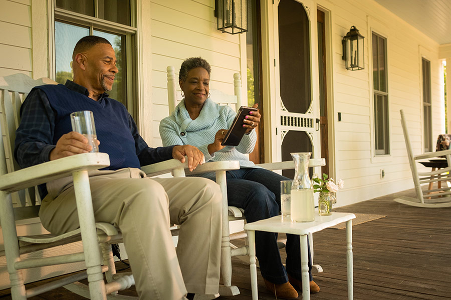 an older couple on the house front porch looking at a tablet, sitting in chairs