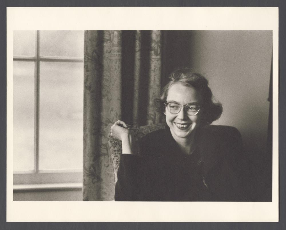 old black and white photo of a woman sitting in a chair smiling, wearing glasses, mid 20th century