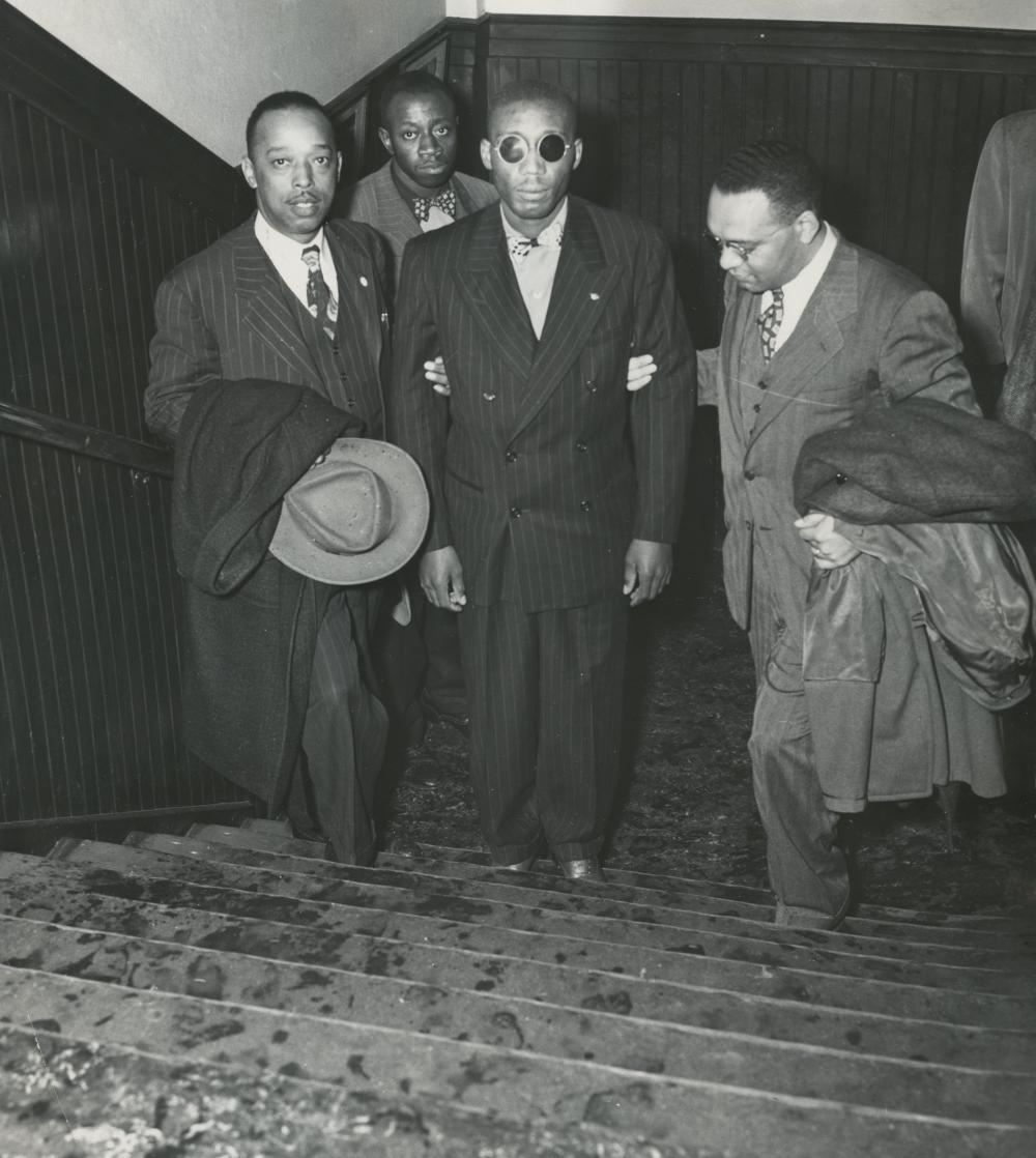 black and white image of men leading another blind man with dark glasses up a staircase. all but the blind man wear suits and carry long coats.